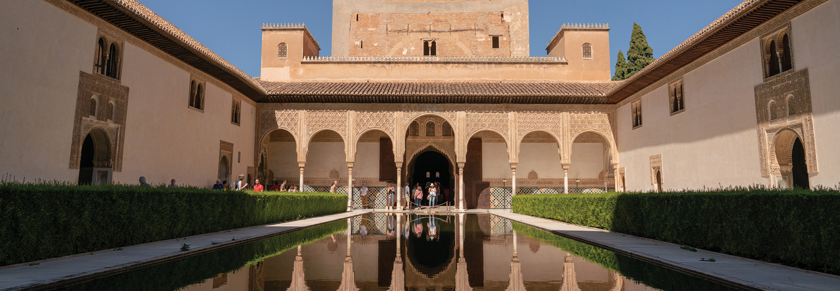 A long reflecting pool mirrors the ornate arches and stucco walls of a sunlit palace courtyard in medieval Spain.