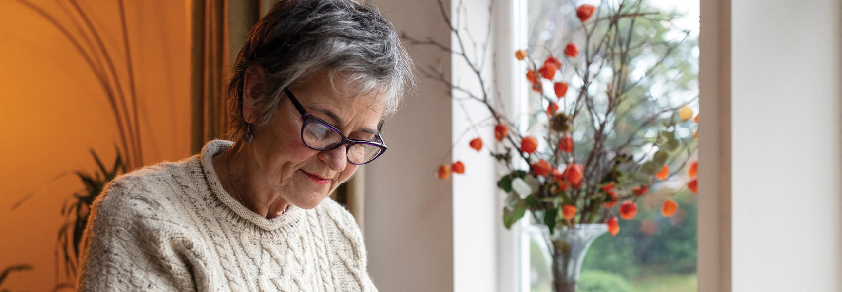 A gray-haired woman wearing glasses and a cable-knit sweater sits by a window, looking down and smiling slightly.
