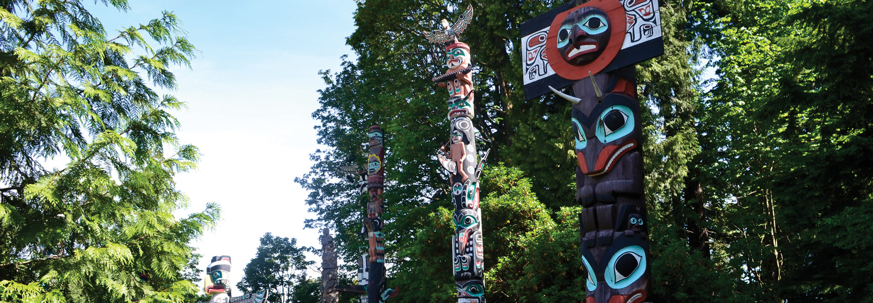 Several colorful First Nations totem poles stand tall among lush green trees under a bright blue sky.