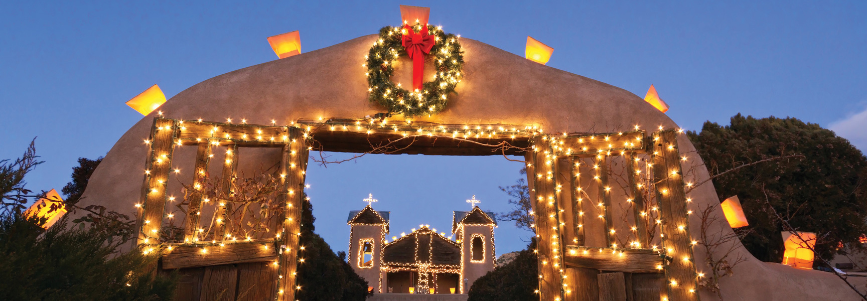 An adobe archway decorated with a Christmas wreath and lights frames a similarly illuminated mission church against a twilight sky.