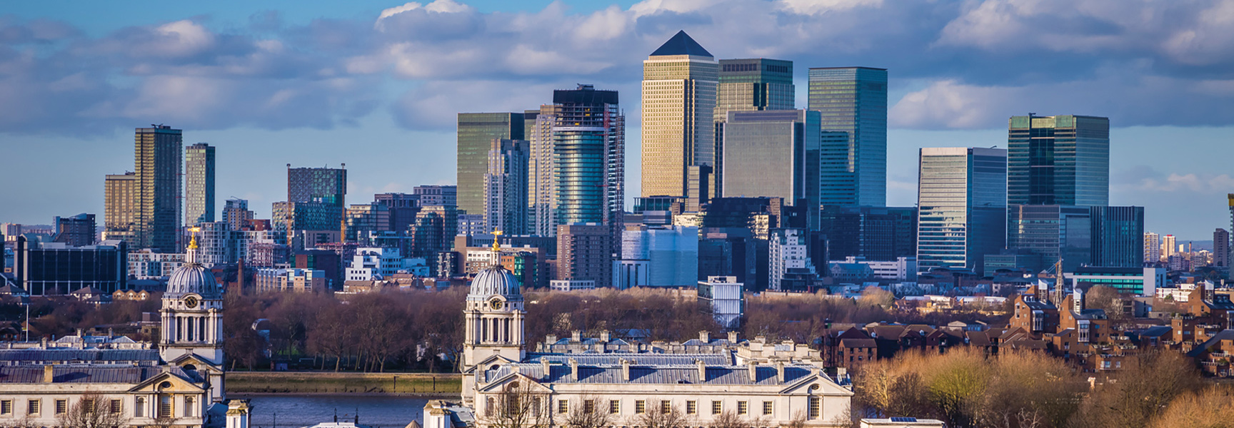 The modern skyscrapers of London, England, rise behind the classical domes of the Old Royal Naval College on a partly cloudy day.