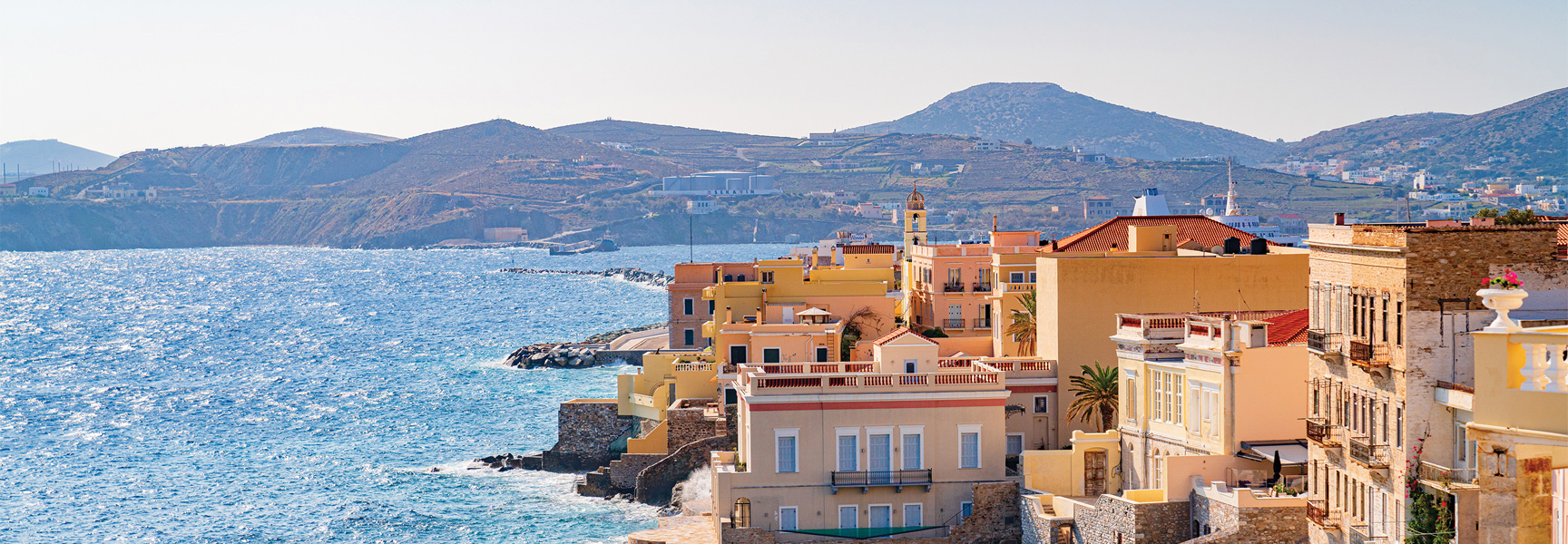 Sunlit, colorful buildings of a coastal town in Greece are nestled against the bright blue sea with arid hills in the distance.