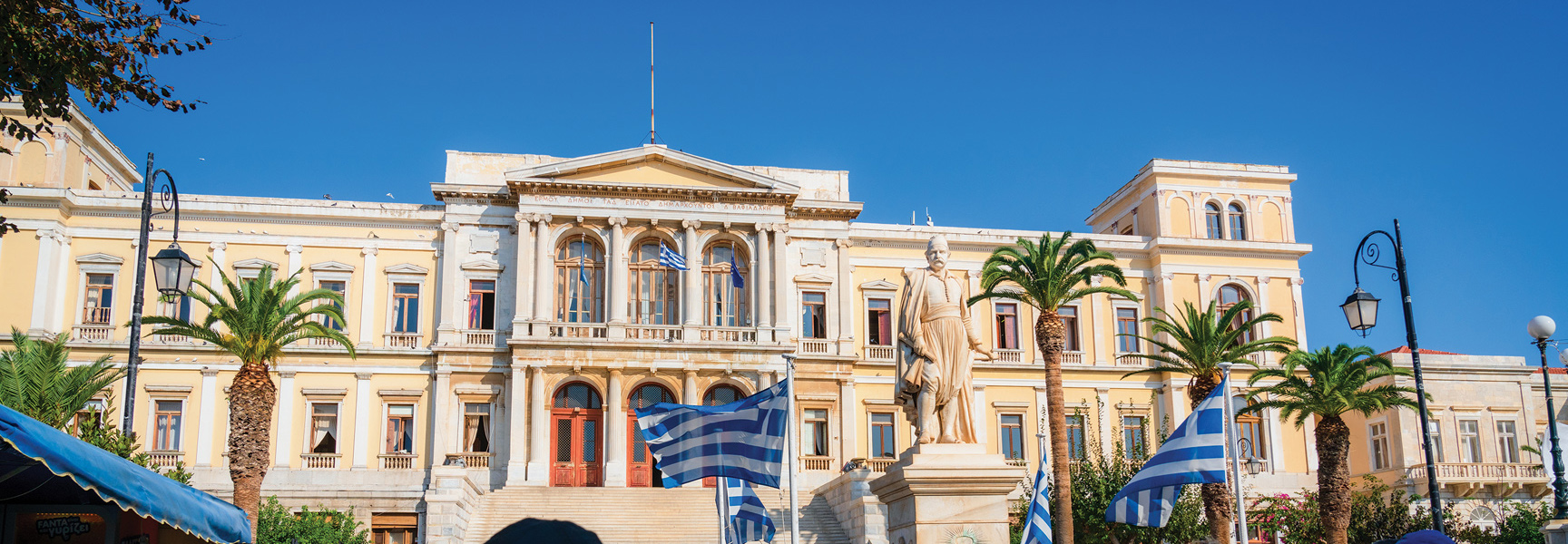 A grand neoclassical building in Greece with a prominent statue and Greek flags out front, framed by palm trees under a sunny sky.