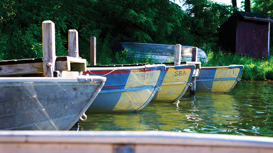 A line of colorful rowboats are tied to a wooden dock on a calm lake with trees in the background.