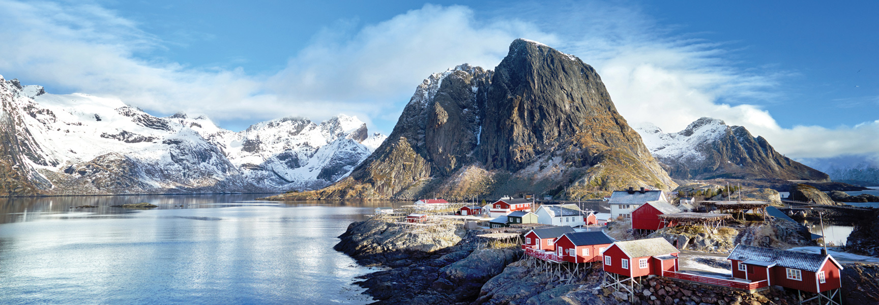 A small coastal village with colorful houses sits at the base of a dramatic, snow-capped mountain range on a fjord in Norway.