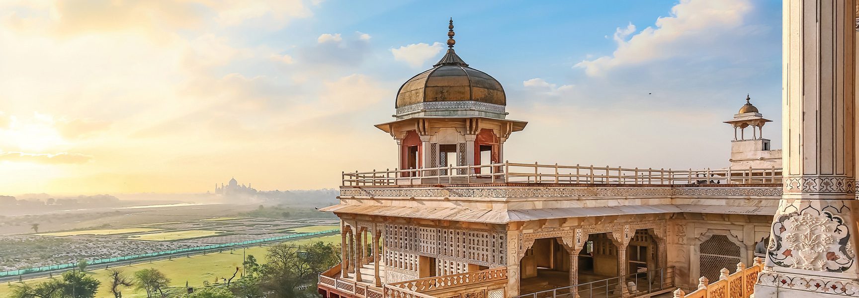An ornate palace balcony in India at sunrise overlooks fields and the distant Taj Mahal, shrouded in morning mist.