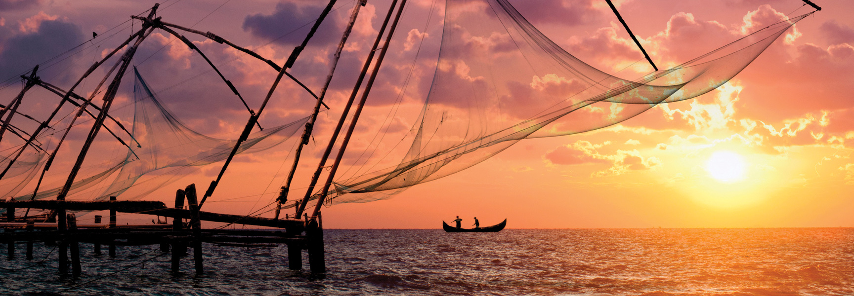 Silhouetted Chinese fishing nets and a small canoe on the water during a brilliant orange and purple sunset.