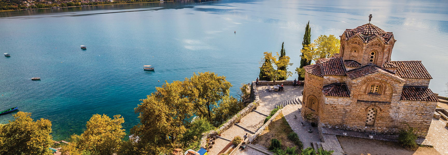 A stone church sits on a cliff overlooking the vast, blue Lake Ohrid in North Macedonia, with boats dotting the water below.