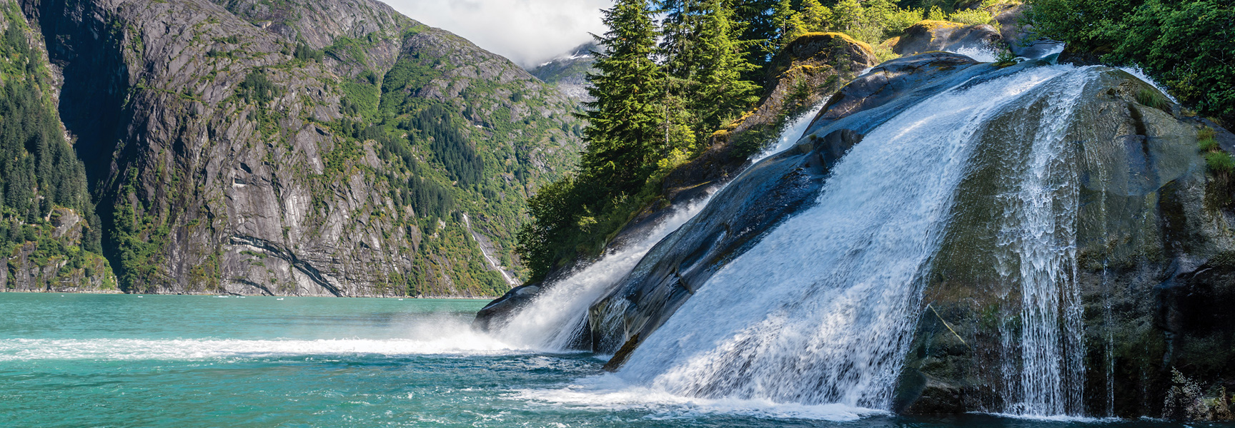 A wide waterfall cascades over a rocky cliff into the turquoise water of a fjord in Alaska, with lush green mountains in the background.