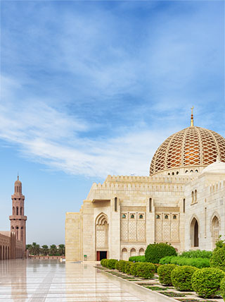 The sunlit courtyard of the Sultan Qaboos Grand Mosque in Oman, with its large dome and minaret under a bright blue sky.