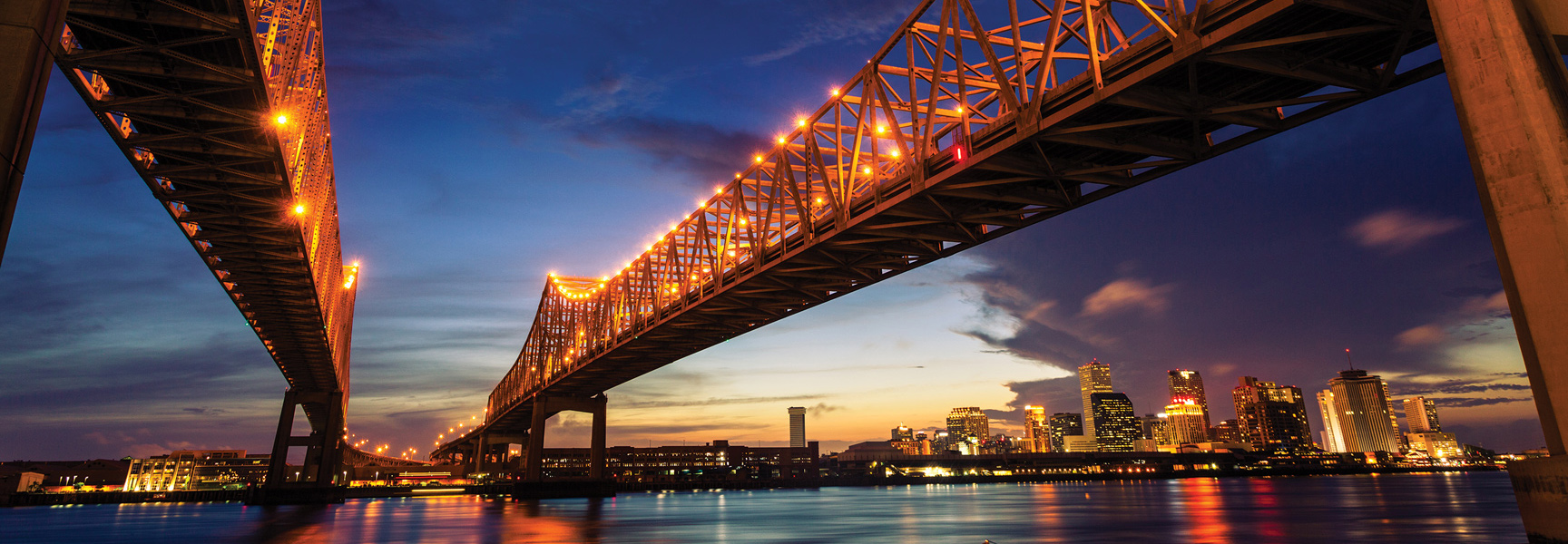 A low-angle view from the Mississippi River shows the illuminated Crescent City Connection bridge at twilight with the New Orleans, Louisiana skyline beyond.