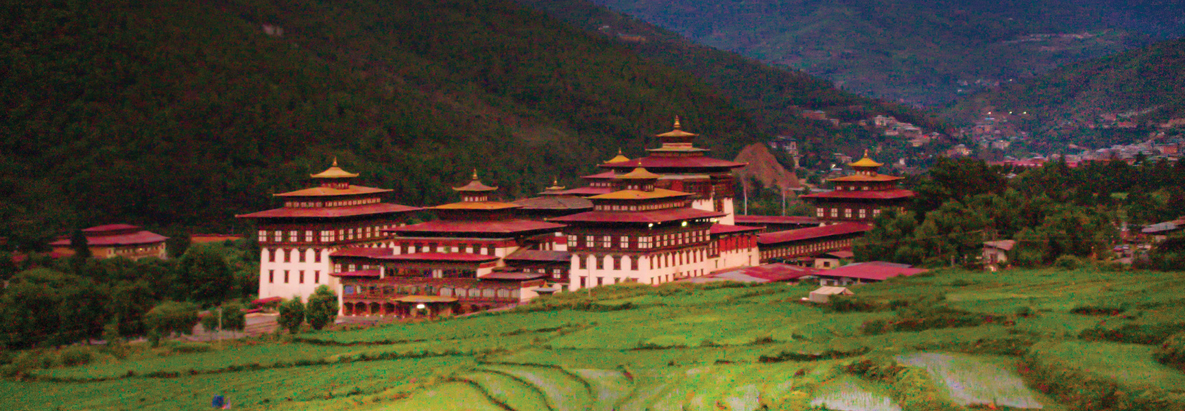A large Bhutanese monastery with red and gold roofs sits in a lush green valley at the base of a forested mountain.