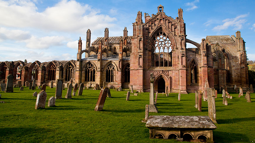 The ruins of Melrose Abbey in Scotland, with its large arched windows and stone facade, stand behind a grassy churchyard filled with weathered tombstones.