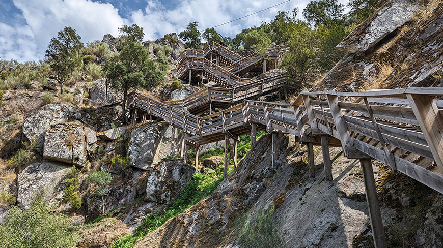 A wooden walkway with many stairs zigzags up a steep, rocky mountainside under a blue sky in Costa Rica.