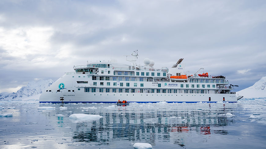 A large white expedition ship, the Greg Mortimer, sails through calm, icy waters with a small boat alongside and mountains behind.