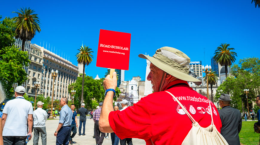 A Road Scholar group leader in a red shirt and sun hat holds up a red sign for their group in a sunny city plaza.