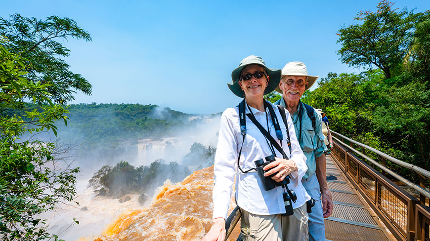 A smiling couple in sun hats stands on a walkway overlooking the powerful, misty Iguazu Falls surrounded by lush jungle.