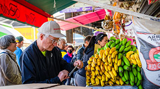 An older man in a baseball cap stands next to a large bunch of bananas at a busy outdoor market, looking down at his phone.