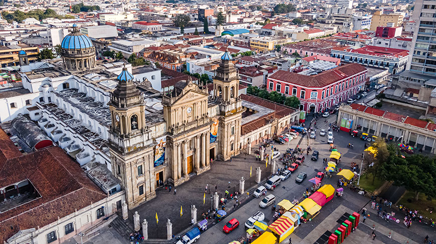 An aerial view of the grand Guatemala City Cathedral next to the bustling Constitution Plaza, with colorful market stalls and city buildings stretching into the distance.