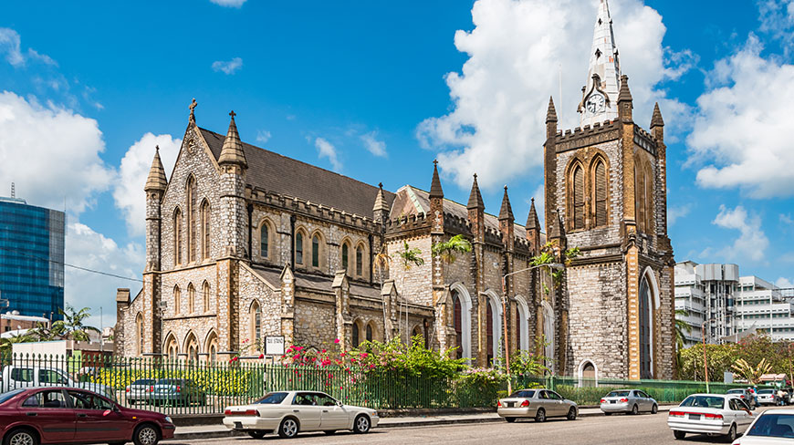 The grey stone Holy Trinity Cathedral in Port of Spain stands under a blue sky, with a city street and cars in the foreground.