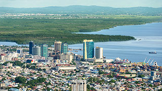 An aerial view of the Port of Spain skyline in Trinidad, with the coast and lush green hills visible in the background.