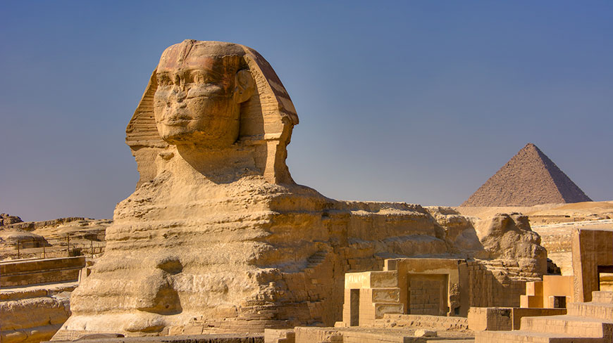 The Great Sphinx of Giza stands in the desert with a large pyramid visible in the background under a clear blue sky.
