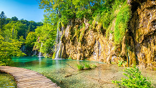 A wooden boardwalk curves alongside a clear turquoise lake with several waterfalls cascading down a lush, green cliffside under a blue sky.