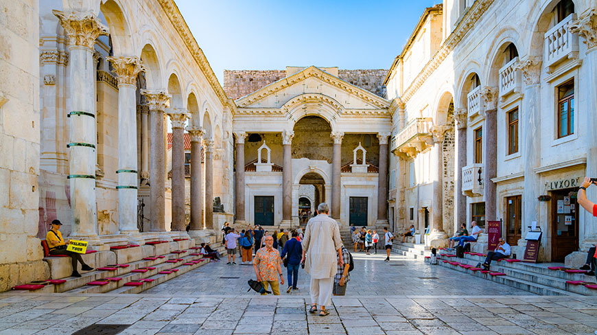 Tourists gather in the sunny, stone courtyard of Diocletian's Palace, surrounded by large Roman columns and archways.
