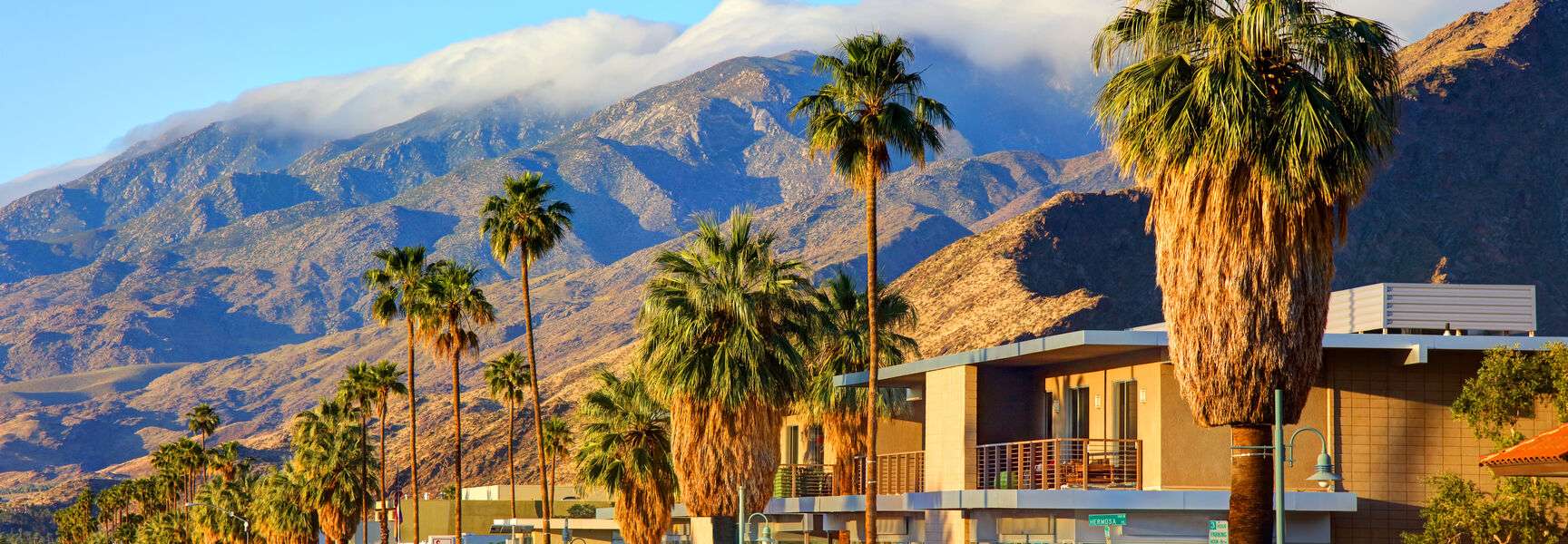 Palm trees line a sunny street in the city of Palm Springs, California, with majestic mountains and clouds in the background.