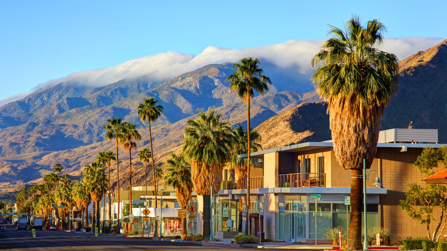 Palm trees line a sunny street in the city of Palm Springs, California, with majestic mountains and clouds in the background.