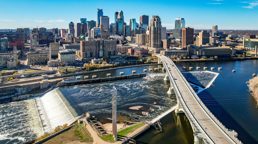 An aerial view of the Minneapolis cityscape featuring the Mississippi River and downtown skyline under a bright blue sky.