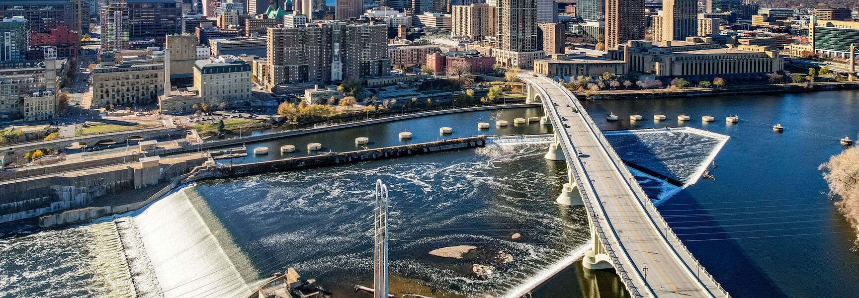 An aerial view of the Minneapolis cityscape featuring the Mississippi River and downtown skyline under a bright blue sky.