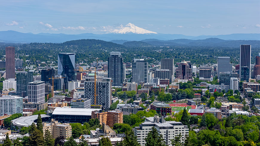 A skyline view of Portland, Oregon, featuring city buildings and the snow-capped Mount Hood in the distance under a clear blue sky.