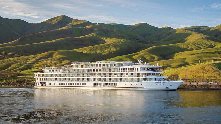 The ACL American Jazz cruise ship afloat on a river with scenic green hills in the background.