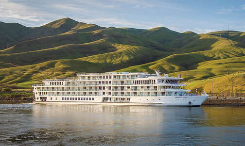 The ACL American Jazz cruise ship afloat on a river with scenic green hills in the background.