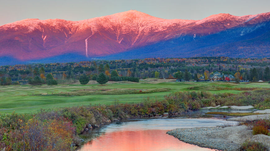 The pink alpenglow on Mount Washington is reflected in a river in Bretton Woods, New Hampshire, during a vibrant autumn sunset.