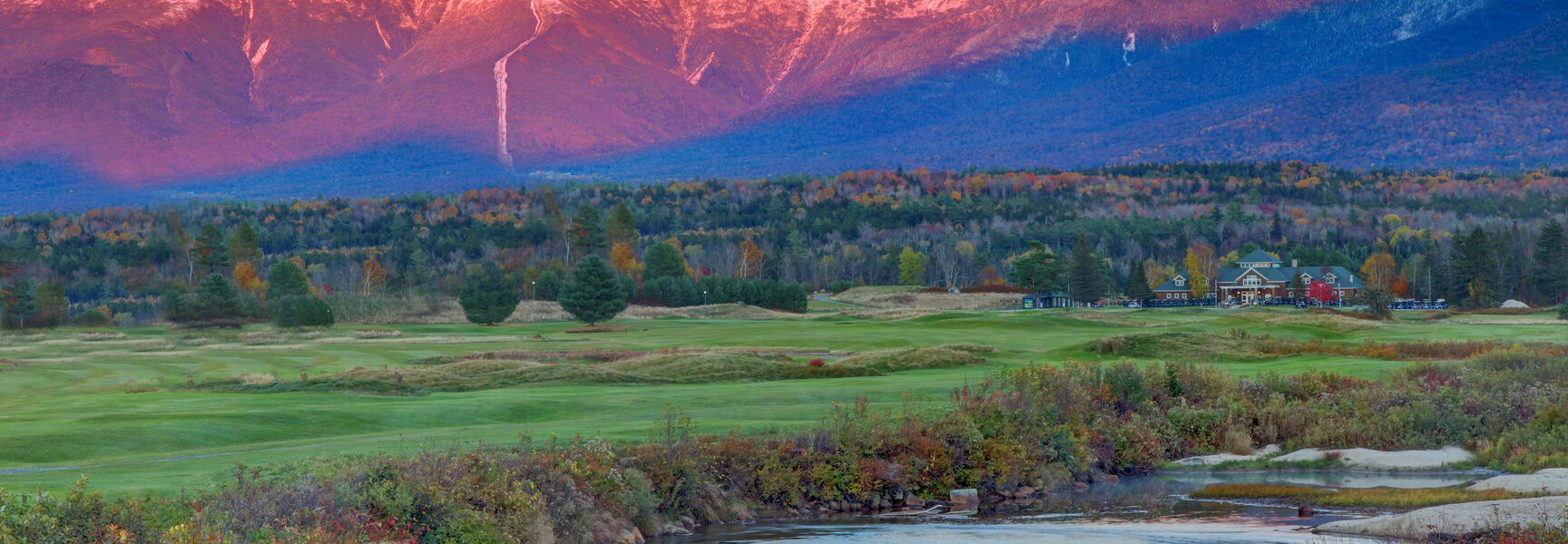 The pink alpenglow on Mount Washington is reflected in a river in Bretton Woods, New Hampshire, during a vibrant autumn sunset.