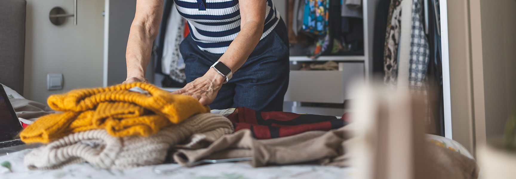 An older woman in a striped shirt folds a pile of clean sweaters on a bed in her bedroom.