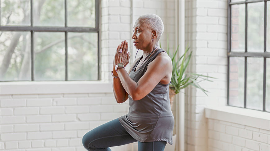 An older Black woman with short gray hair holds a yoga pose in a sunlit room with large windows.