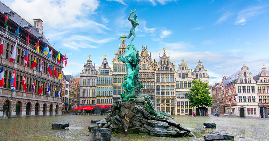 The Brabo Fountain in Antwerp's Grote Markt, surrounded by historic guildhouses and the city hall decorated with colorful flags under a bright sky.