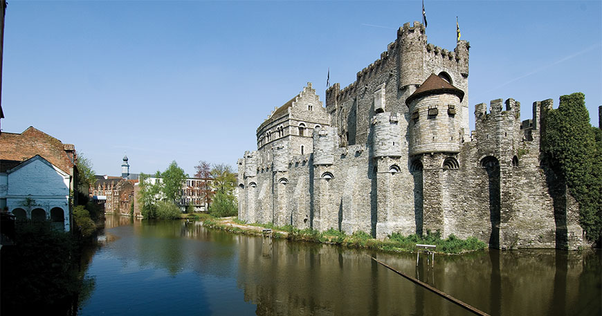A medieval stone castle with turrets and flags stands along a canal in Ghent, Belgium, under a clear blue sky.