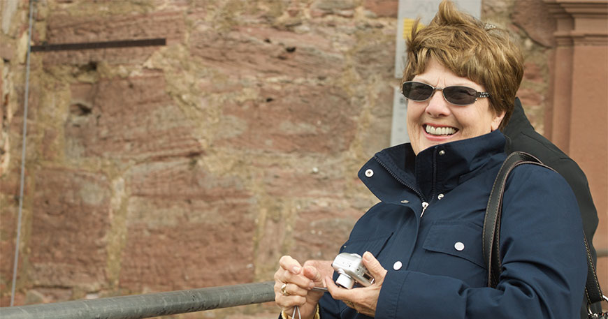 A smiling woman wearing a navy blue jacket and sunglasses holds a silver digital camera while standing in front of a stone wall.
