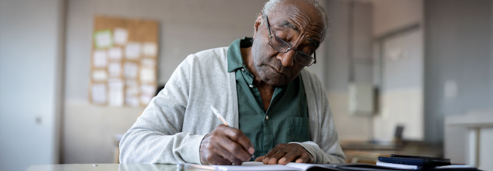 An older man with glasses wearing a cardigan sits at a desk and writes in a notebook with a pen.