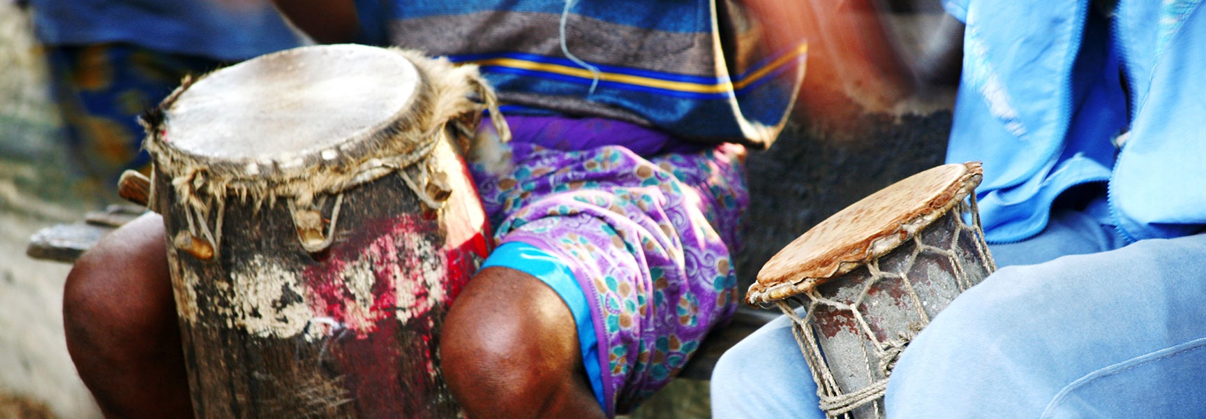 A close-up of two African drummers sitting with traditional talking drums held between their knees.