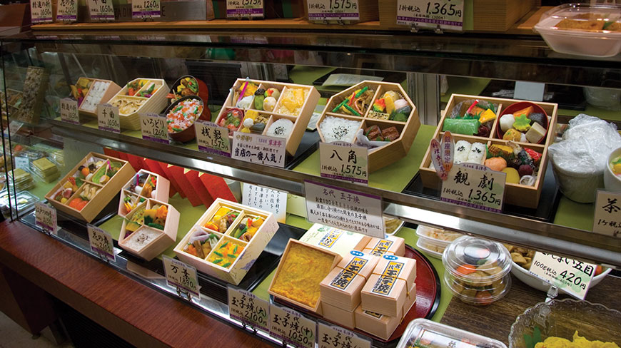 A variety of Japanese bento boxes with colorful food arrangements are displayed for sale inside a glass case.