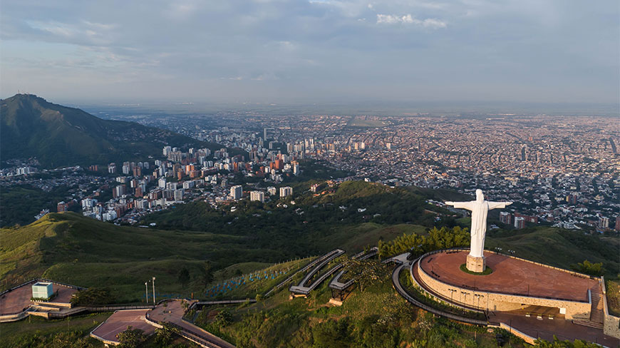 The white Cristo Rey statue overlooks the expansive city of Cali, Colombia, from a hilltop plaza in this wide aerial view.