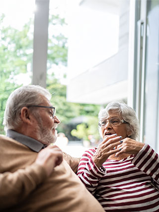 An older man and woman with glasses sit on a couch and have an animated conversation.