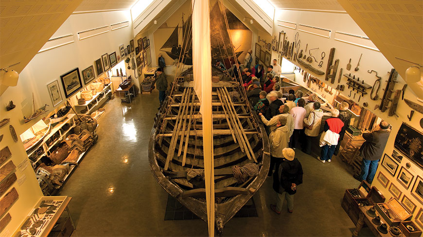 Visitors gather around a large wooden boat displayed at the Skogar Museum, surrounded by various maritime artifacts on the museum walls.