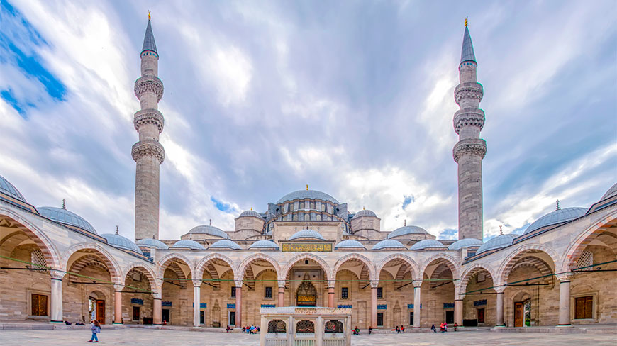 Wide-angle view of a grand mosque courtyard in Istanbul featuring arched walkways, multiple domes, and two towering stone minarets under a cloudy sky.