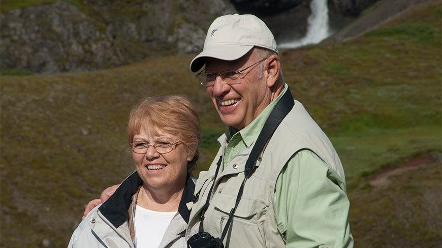 A smiling couple, the Petersons, stand together outdoors with a waterfall and rolling hills in the background.